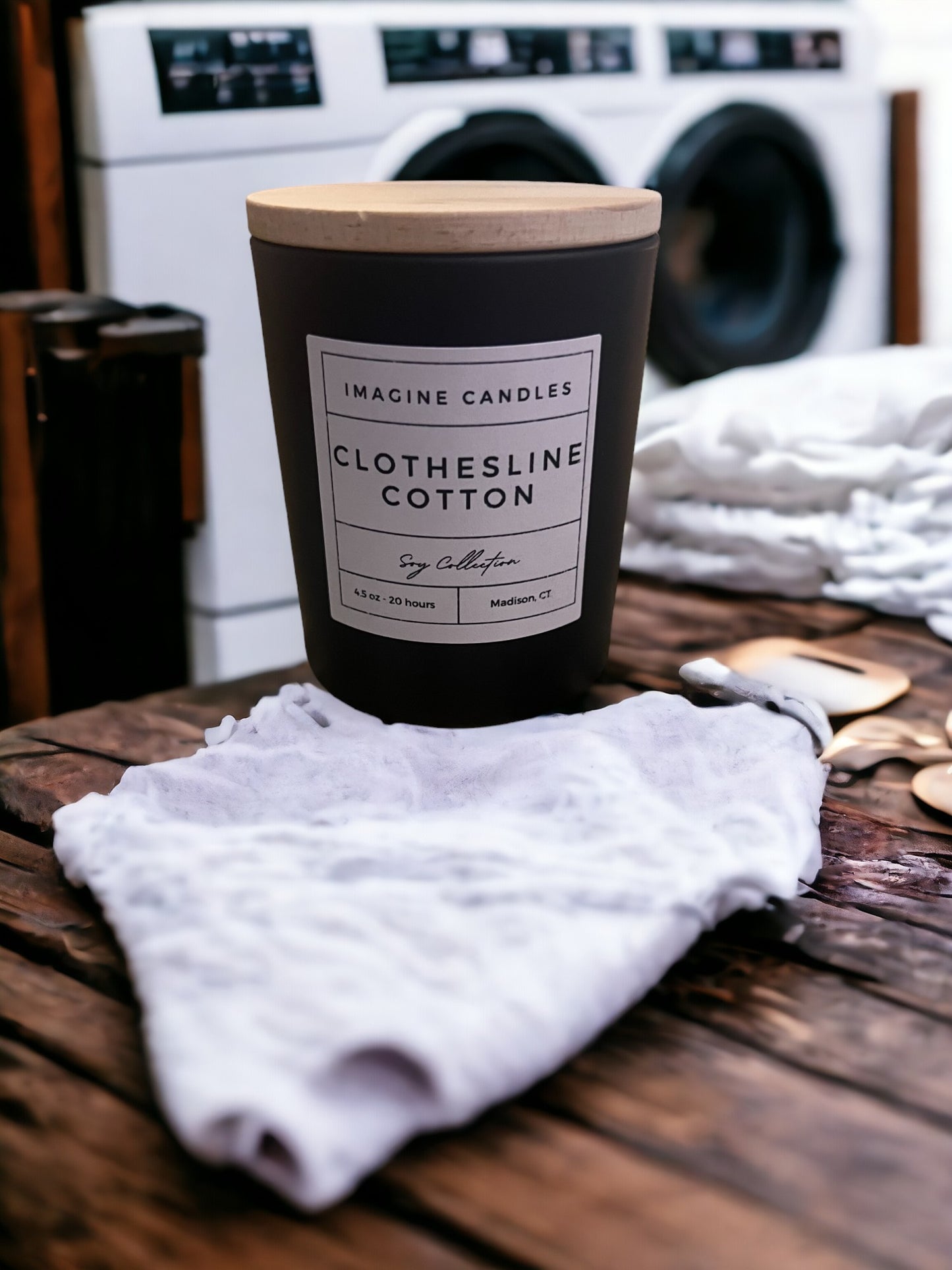 A candle sits on a clean countertop in a tidy laundry room. The label reads 'Clothesline Cotton', evoking the scent of freshly cleaned laundry. The fragrance notes include ozone, powder, cotton blossom, lily of the valley, light musk, and violet. Infused with natural essential oils, including lemon peel