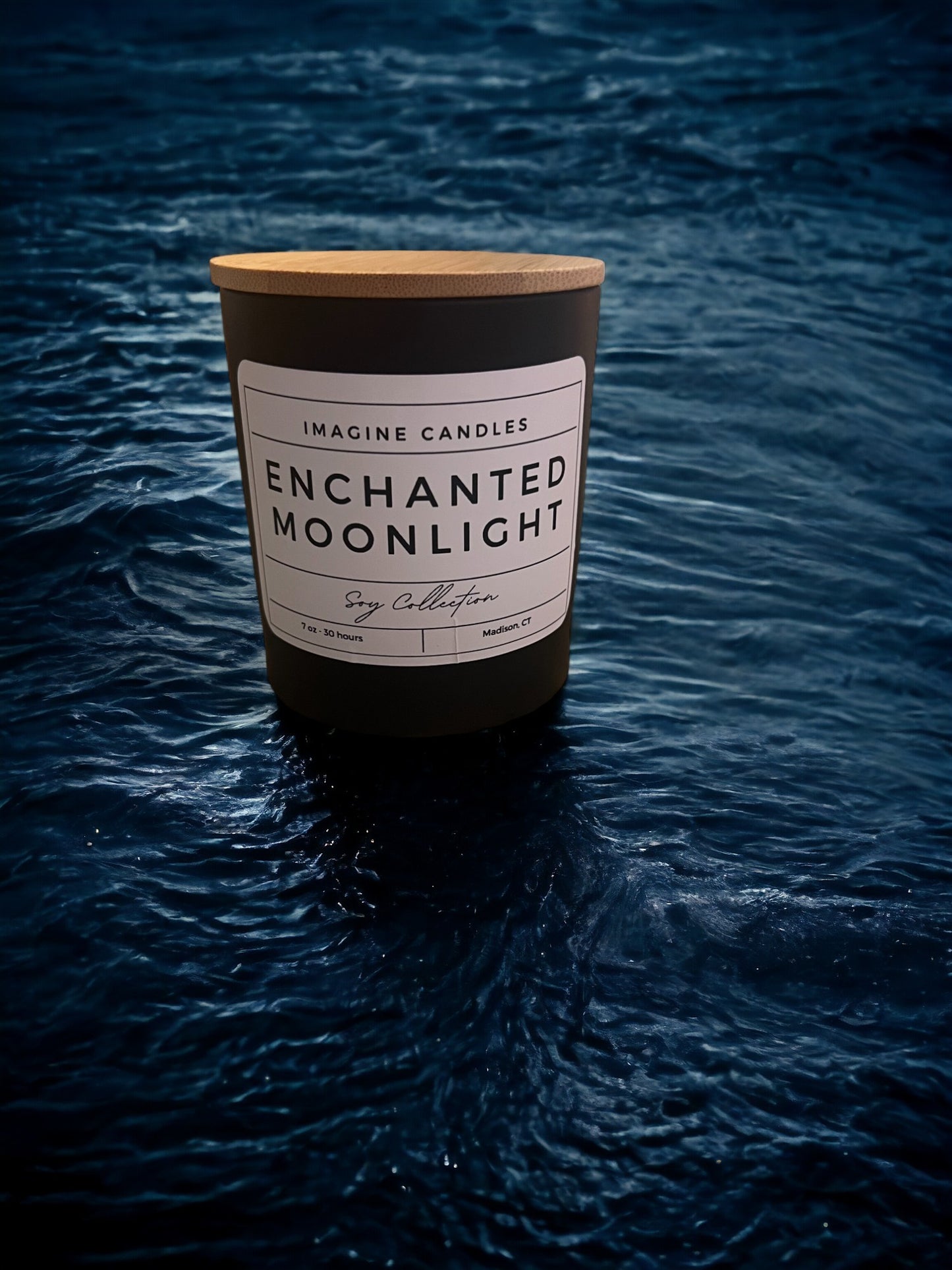 A candle rests overlooking moonlit water. The label reads 'Enchanted Moonlight', evoking the serene ambiance of a starry night. The fragrance notes include bergamot, citrus, green, aromatic, spice, herbal, cedarwood, and sandalwood