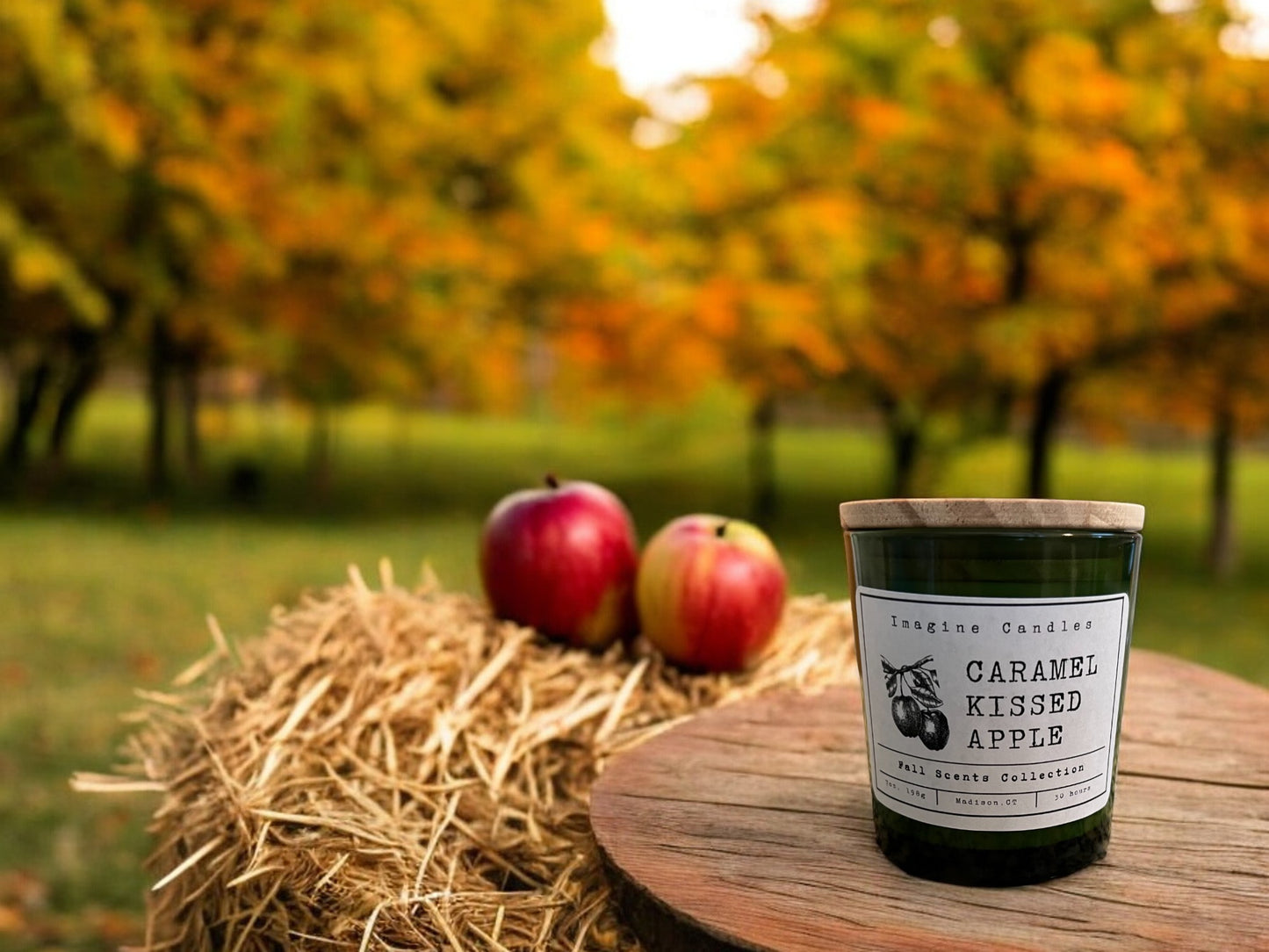 A green candle jar sits on a hay bale with two apples in the trees in the background are changing colors