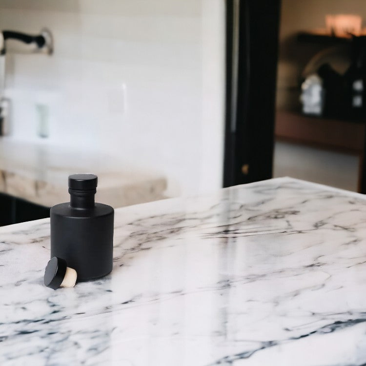 A black reed diffuser vessel sits on a marble countertop. The description introduces the Matte Black Round Reed Diffuser Bottle, emphasizing its sleek and sophisticated design. With a velvety matte finish and timeless noir hue, the vessel exudes understated elegance, suitable for luxury and minimalist home fragrance lines. Crafted with meticulous attention to detail, it offers a modern appeal that seamlessly complements any decor style, adding sophistication to any room.