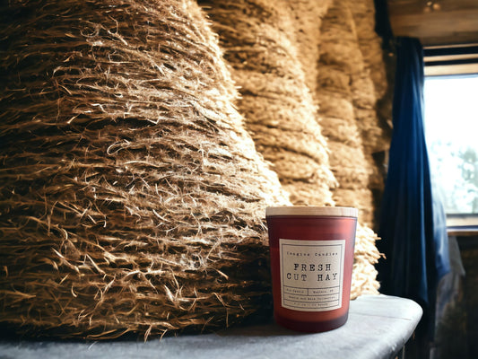 A candle sits amidst a stack of fresh hay, emitting the scent of the outdoors. The label reads 'Fresh Cut Hay', capturing the essence of new mown hay with sweet balsam, vetiver, and cedarwood notes. A delightful fragrance reminiscent of fresh country air
