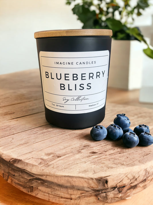 A Blueberry Bliss candle sits on a wooden counter next to fresh blueberries, embodying the essence of indulgence. Fragrance notes include butter, sugar, blueberry, graham cracker, and vanilla, reminiscent of a once-a-year blueberry pie from the local farmer’s market.