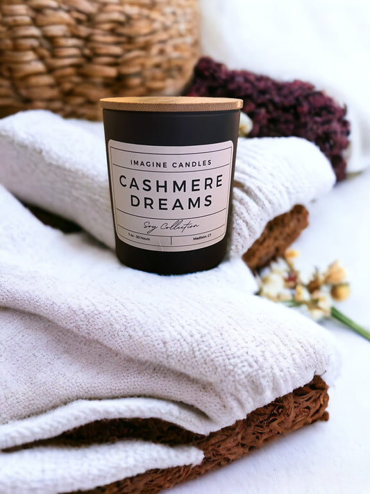 A candle rests atop a stack of neatly folded towels. The label reads 'Cashmere Dreams', evoking the sensation of comfort and relaxation. The fragrance notes include bergamot, saffron, violet, amber, lily of the valley, dark musk, vetiver, and powder.
