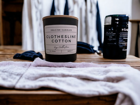 A candle sits on a clean countertop in a tidy laundry room. The label reads 'Clothesline Cotton', evoking the scent of freshly cleaned laundry. The fragrance notes include ozone, powder, cotton blossom, lily of the valley, light musk, and violet. Infused with natural essential oils, including lemon peel