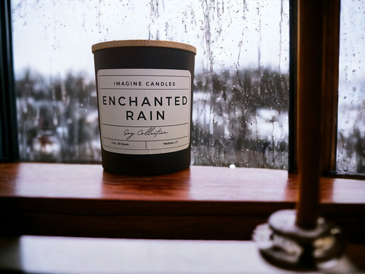A candle sits on a rainy windowsill, evoking the ambiance of the first rainfall of Spring. The label reads 'Enchanted Rain', capturing the essence of the purifying rain. The fragrance notes include green leaves, lily of the valley, rose, ozone, and sandalwood. Infused with natural essential oils, including orange peel, lemon peel, and petitgrain leaf