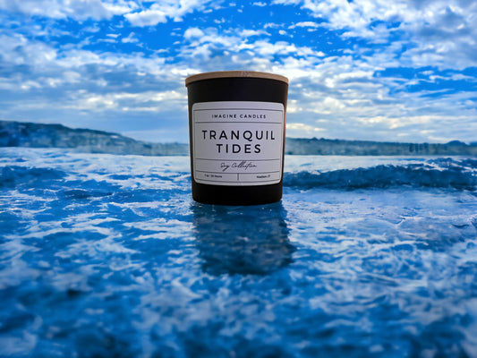 A candle rests on blue ocean waves, embodying the essence of Tranquil Tides. The label reads 'Tranquil Tides', evoking the feeling of the first day of a beach vacation, relaxing by an open window. Fragrance notes include bergamot, sea salt, peony, mint, juniper, light musk, and marine, infused with natural essential oils, including lemon, peppermint, and lime.