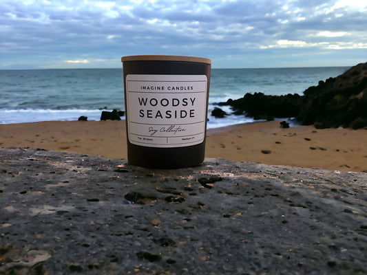 A candle rests at the edge of the woods and beach, epitomizing the essence of Woodsy Seaside. The label reads 'Woodsy Seaside', evoking the symbiosis of yin and yang where forest meets ocean. Fragrance notes include sea salt, orange peel, grass, eucalyptus, sage, marine, patchouli, teakwood, and moss, infused with natural essential oils, including patchouli, cedarwood, clove leaf, elemi, eucalyptus, orange sweet, guaiacwood, cedar, pine, clary sage, and galbanum