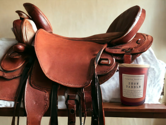 A candle sits in front of a western show saddle, evoking the scent of Show Saddle. The label reads 'Show Saddle', capturing the feeling of pre-class jitters and the comforting squeak of tacking up. Fragrance notes include saffron, incense, leather, and amber